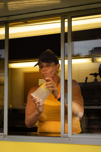 A woman in a yellow tank top and black cap shakes a cup of lemonade.